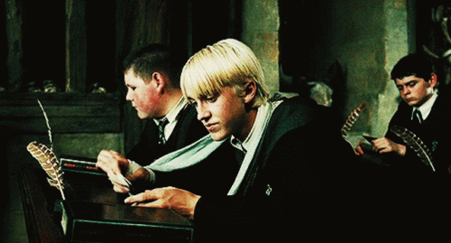 A Group Of Young Men Are Sitting At A Table With Feathers And A Book That Says Harry Potter On It GIF