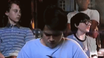 A Group Of Young Men Are Sitting In A Dark Room . One Of The Men Is Wearing Glasses And A Blue Shirt . GIF