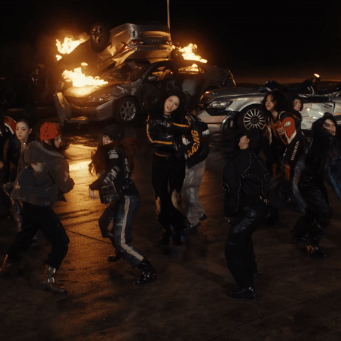A Group Of Young Women Are Dancing In Front Of A Pile Of Burning Cars GIF
