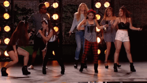 A Group Of Young Women Are Dancing On A Stage With A Man Holding A Microphone GIF
