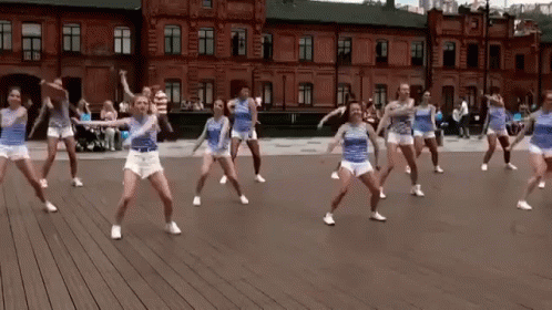 A Group Of Young Women Are Dancing On A Wooden Floor In Front Of A Brick Building . GIF