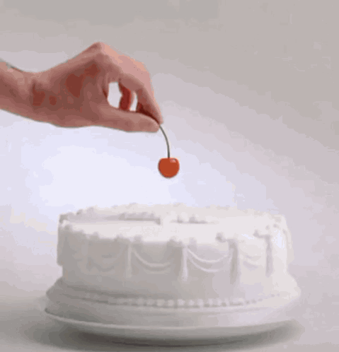 A Hand Holds A Cherry Over A White Cake GIF