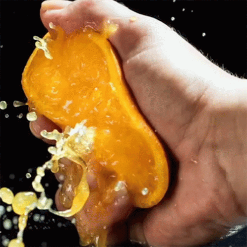 A Hand Is Squeezing A Slice Of Orange With Juice Splashing Out Of It GIF