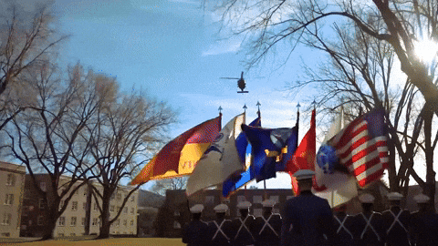 a helicopter flies over a line of soldiers holding flags including one that says ' army ' on it