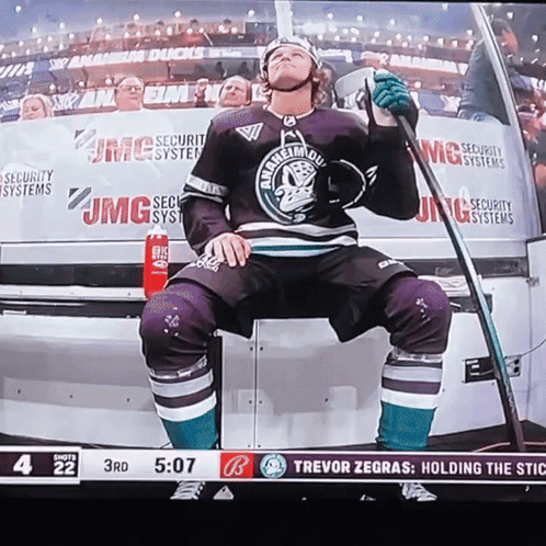 A Hockey Player Is Sitting On The Bench Holding A Stick With Jmg Security Systems In The Background GIF