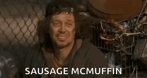 A Homeless Man Is Sitting In Front Of A Chain Link Fence With The Words `` Sausage Mcmuffin '' . GIF