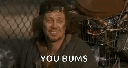 A Homeless Man Is Sitting In Front Of A Chain Link Fence With The Words `` You Bums '' Written On His Face . GIF