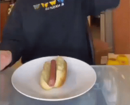 A Hot Dog On A Plate With A Person Behind It GIF