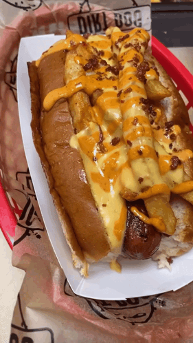A Hot Dog With Cheese And French Fries Is In A Paper Container GIF