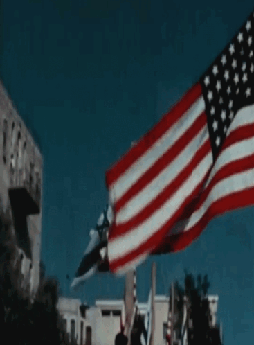 A Large American Flag Is Flying In The Wind In Front Of A Building GIF