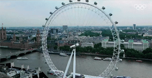 A Large Ferris Wheel In The Middle Of A City With The Olympics Logo On The Bottom GIF