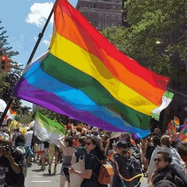 A Large Rainbow Flag Is Being Flown In A Crowded Street GIF