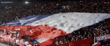 A Large Red , White And Blue Flag Is Being Waved By A Crowd Of People In A Stadium . GIF