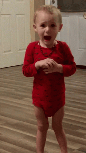 A Little Boy In A Red Bodysuit Is Standing On A Wood Floor GIF