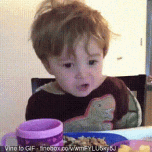 A Little Boy Is Sitting At A Table With A Plate Of Food And A Pink Cup GIF