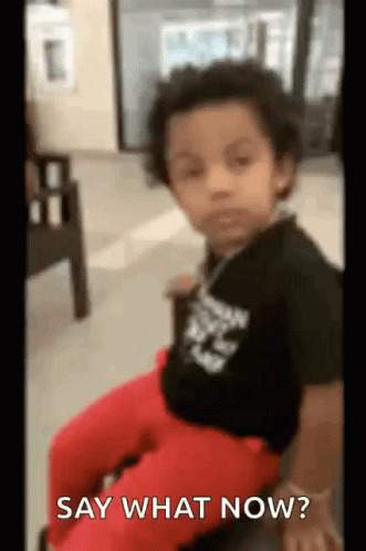 A Little Boy Is Sitting In A Chair With The Words `` Say What Now '' Written On The Screen . GIF