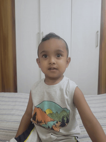 A Little Boy Is Sitting On A Bed Wearing A White Tank Top With Mountains On It GIF