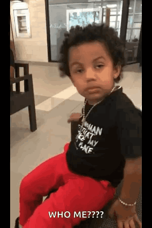 A Little Boy Is Sitting On A Chair And Looking At The Camera While Wearing A Black Shirt And Red Pants . GIF
