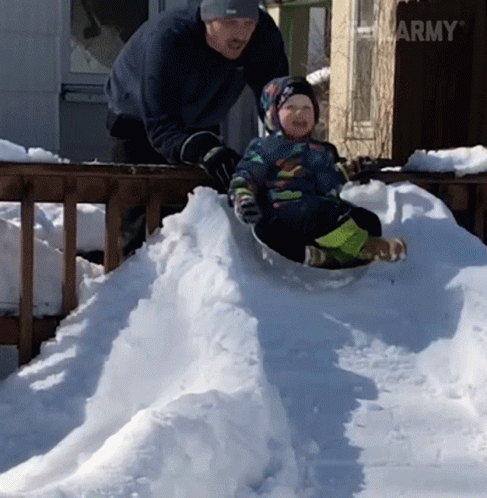 A Little Boy Is Sliding Down A Snowy Hill While A Man Stands Behind Him GIF