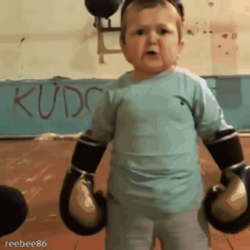 A Little Boy Wearing Boxing Gloves Is Standing In Front Of A Wall That Says Kudc GIF