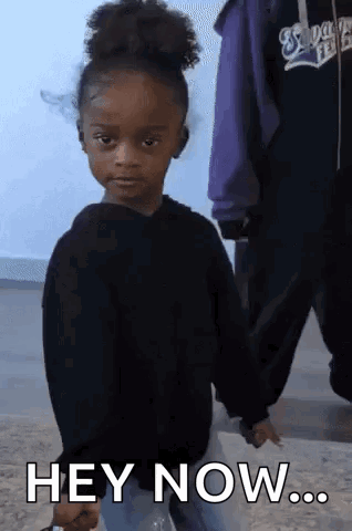 a little girl in a black sweater is holding a bottle and says hey now