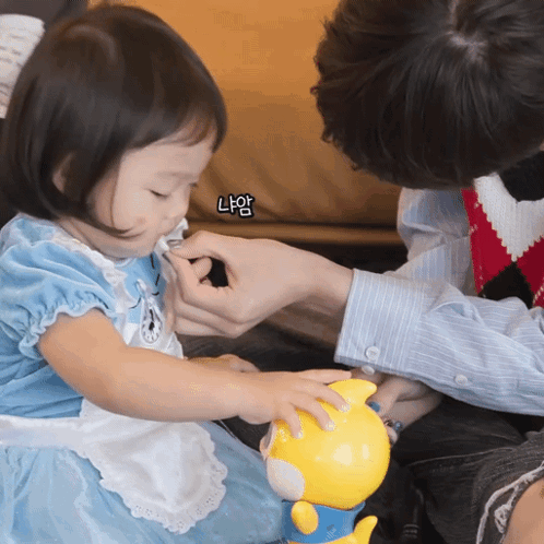 A Little Girl In A Blue Dress Is Playing With A Yellow Toy While A Man Holds Her Hand GIF