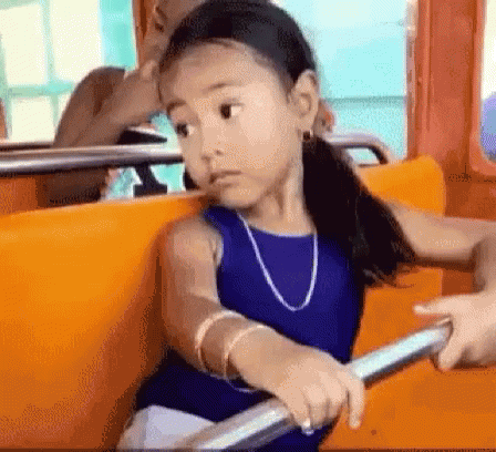A Little Girl In A Blue Dress Is Sitting On A Bus Holding Onto A Railing . GIF