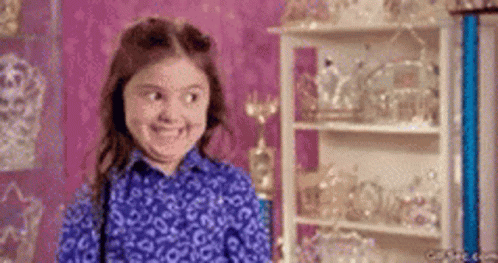 A Little Girl In A Blue Shirt Is Smiling In Front Of A Shelf Full Of Trophies . GIF