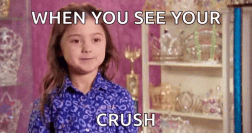 A Little Girl In A Blue Shirt Is Smiling While Standing In Front Of A Shelf With Trophies On It . GIF
