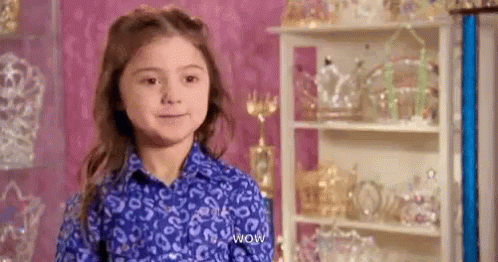 A Little Girl In A Blue Shirt Is Standing In Front Of A Shelf Full Of Trophies . GIF