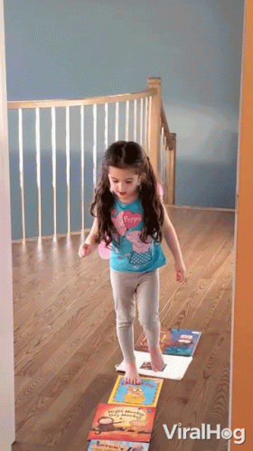 A Little Girl In A Peppa Pig Shirt Is Standing On A Stack Of Books On The Floor GIF