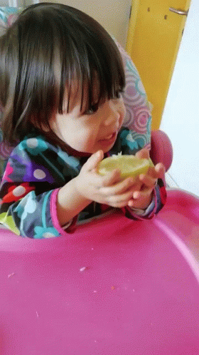 A Little Girl In A Pink High Chair Is Eating A Lemon GIF