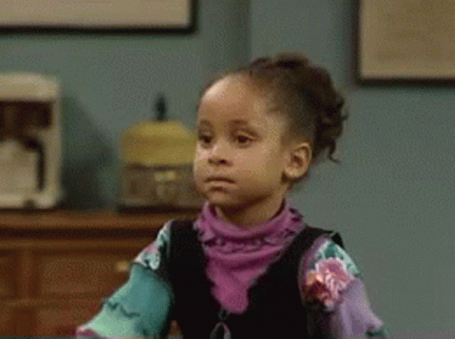 A Little Girl In A Purple Shirt Is Sitting At A Table Making A Funny Face . GIF