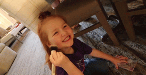 A Little Girl In A Purple Shirt Is Smiling While Holding A Brush In Her Hand GIF