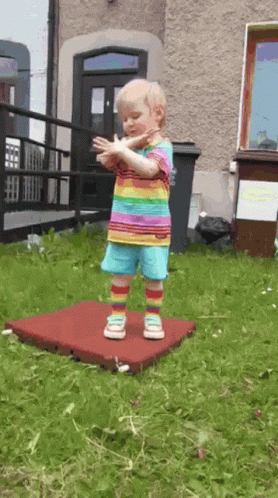 A Little Girl In A Rainbow Shirt And Shorts Is Standing On A Red Mat GIF