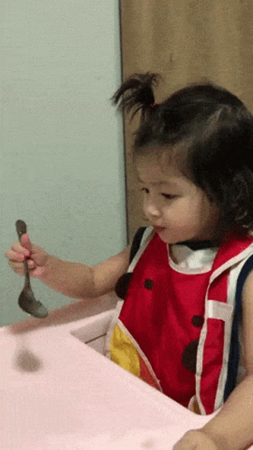 A Little Girl In A Red And White Bib Is Holding A Spoon GIF