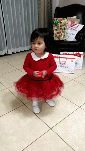 A Little Girl In A Red Dress Is Standing In Front Of A Chair And Christmas Presents GIF