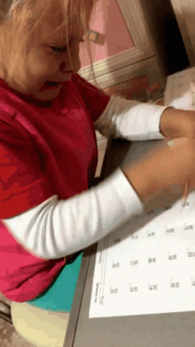 A Little Girl In A Red Shirt Is Sitting At A Desk With A Calendar On It GIF