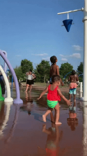 A Little Girl In A Red Top And Green Shorts Is Playing In A Water Fountain GIF