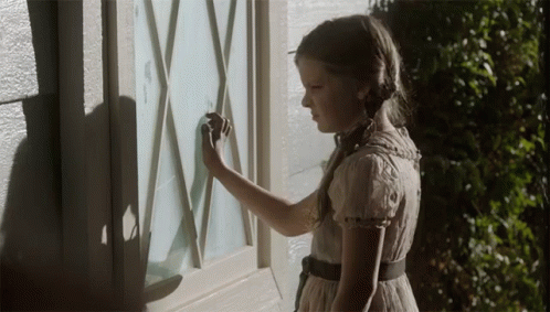 A Little Girl In A White Dress Is Looking At A Window GIF