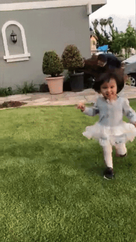 A Little Girl In A White Dress Is Running On The Grass In Front Of A House GIF