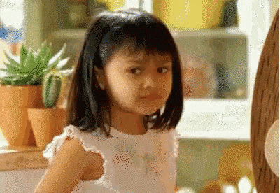 A Little Girl In A White Dress Is Standing In A Kitchen Looking At The Camera . GIF