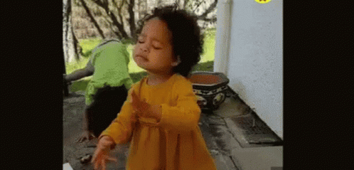 A Little Girl In A Yellow Dress Is Dancing In Front Of A Potted Plant And A Man In A Green Shirt . GIF