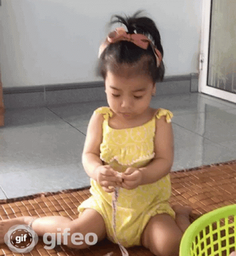 A Little Girl In A Yellow Dress Is Sitting On The Floor Playing With A Ribbon And A Basket With Gif Written On It GIF