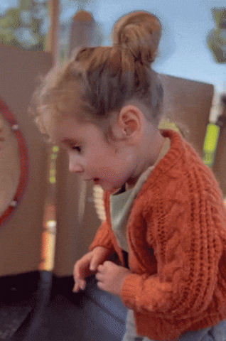 A Little Girl In An Orange Sweater Is Playing With Her Hands GIF