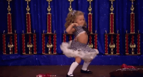 A Little Girl Is Dancing On A Stage With Trophies Behind Her GIF