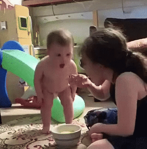 A Little Girl Is Feeding A Baby From A Bowl On The Floor GIF