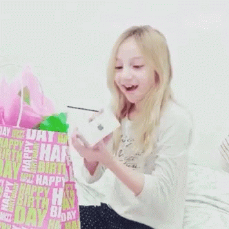 A Little Girl Is Holding A Box In Front Of A Birthday Bag That Says Happy Birthday GIF