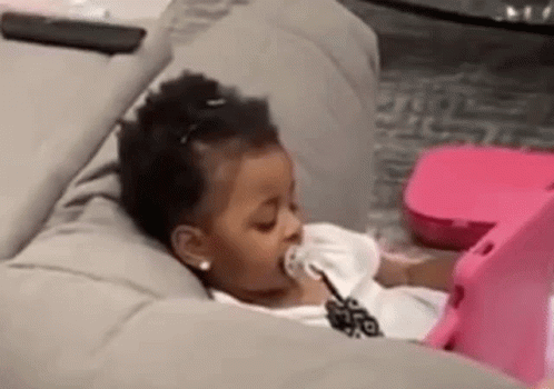 A Little Girl Is Laying On A Couch With A Pacifier In Her Mouth And Looking At A Tablet . GIF