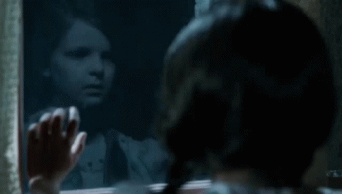 A Little Girl Is Looking Out A Window At Her Reflection In The Mirror . GIF